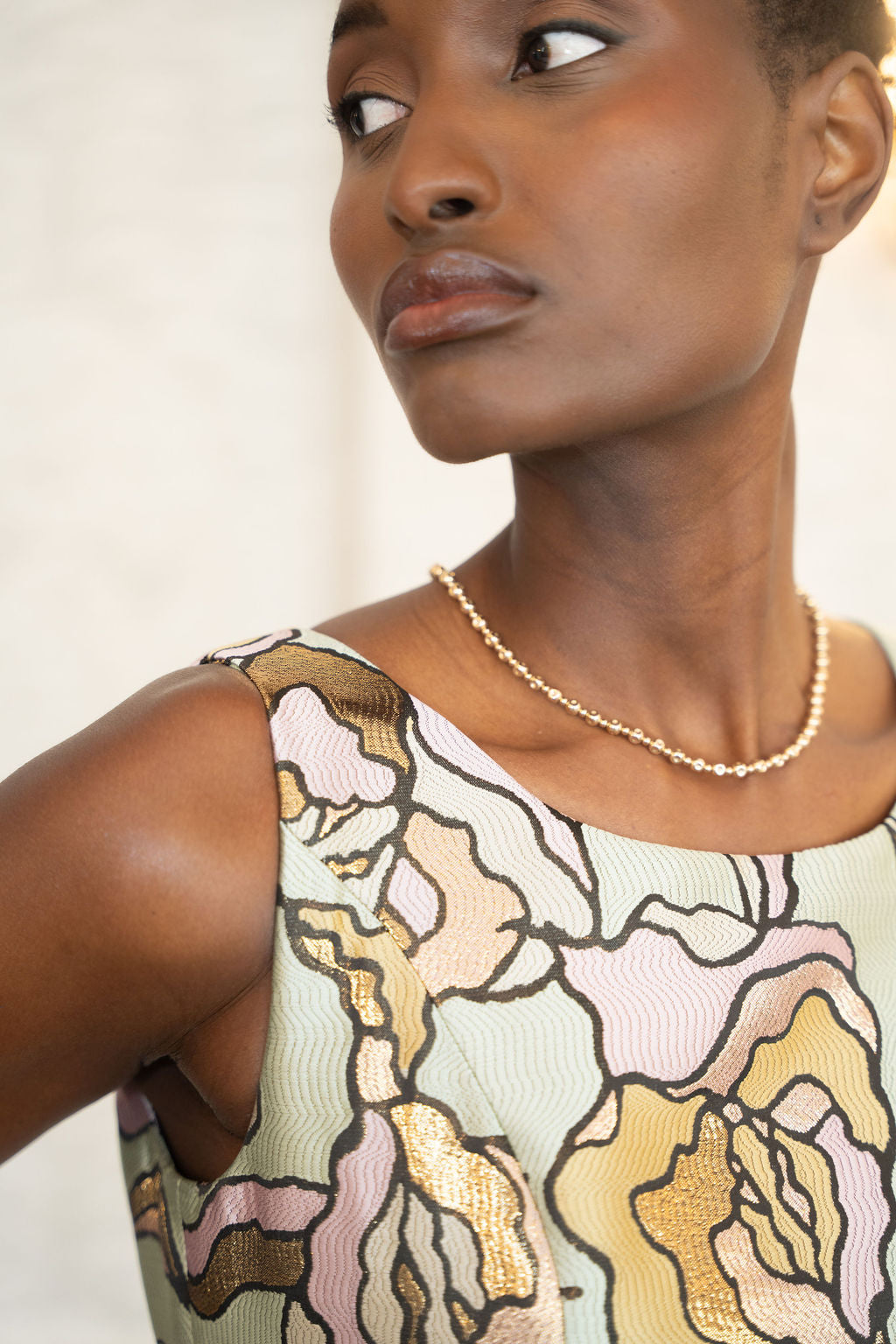 Sleeveless floral brocade midi dress with pastel yellow, pink, and green pattern, worn with a pearl necklace.