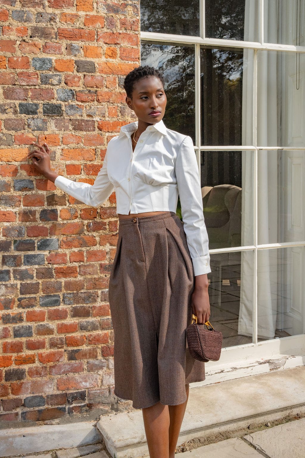 Ivory cotton corset shirt with long sleeves paired with high-waist brown wide-leg trousers and small brown handbag.