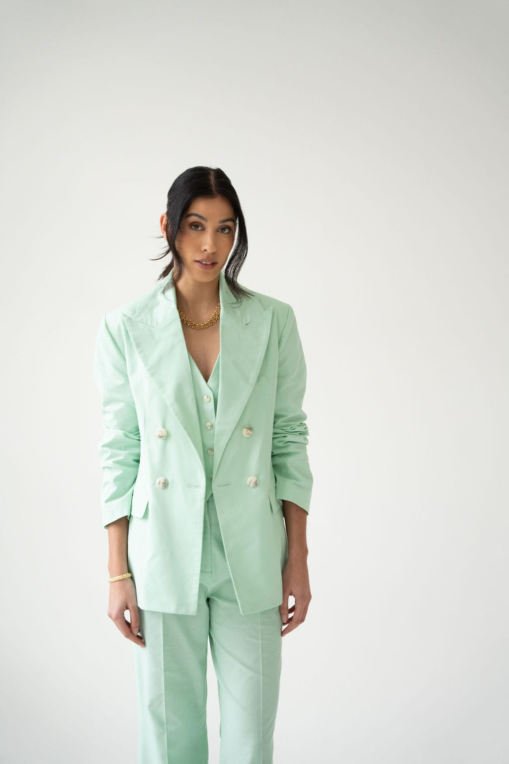 A woman stands against a plain white background wearing the Atelier Ferrari+Monti Double-Breasted Blazer in light mint green cotton, paired with a coordinating vest. Embracing women’s cotton suiting, she styles her long dark hair loosely and accessorizes with gold jewelry, effortlessly blending elegance and comfort.