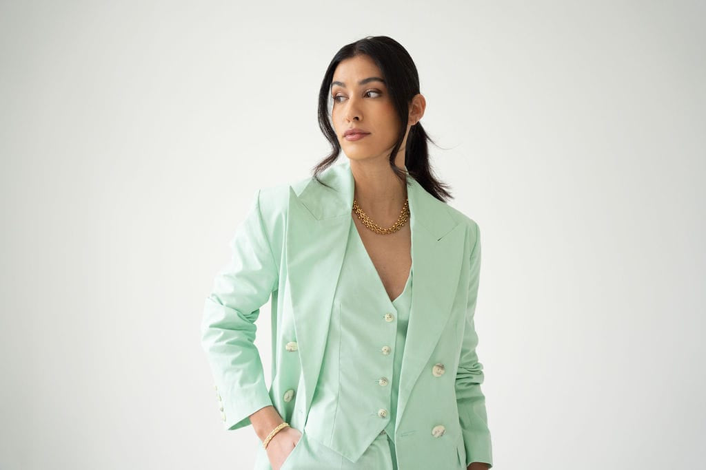 A woman with long dark hair wears the Atelier Ferrari+Monti Double-Breasted Blazer in an oversized mint green cotton style, paired with a gold necklace. She stands against a plain white background, looking to her left with a neutral expression.