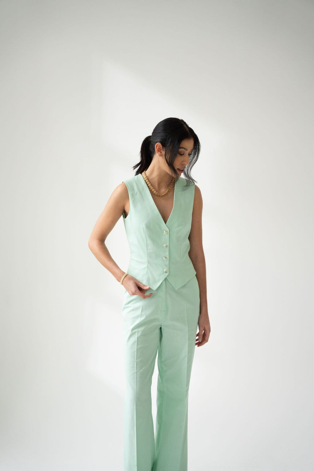 A person stands against a plain white background, looking down with their hair pulled back, wearing Atelier Ferrari+Monti Wide-Leg Tailored Trousers and a matching sleeveless mint green cotton vest.