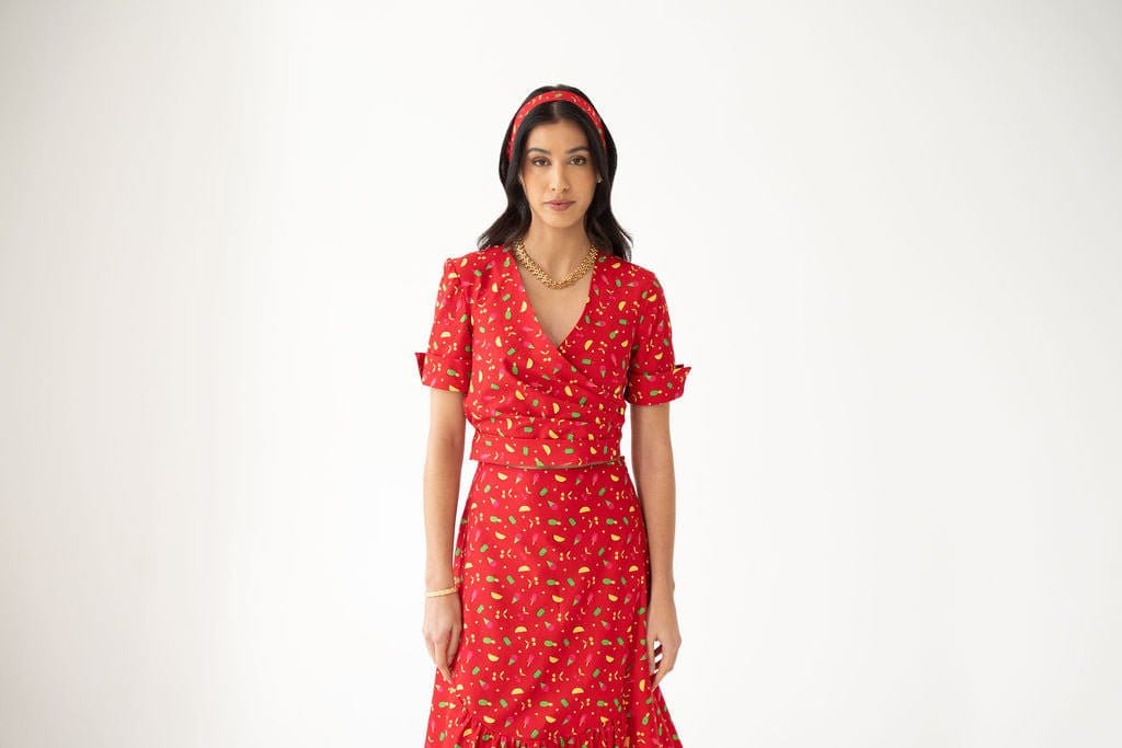 A woman wearing an Atelier Ferrari+Monti Gelato Print Wrap Top, matching skirt, and red headband stands against a plain white background.
