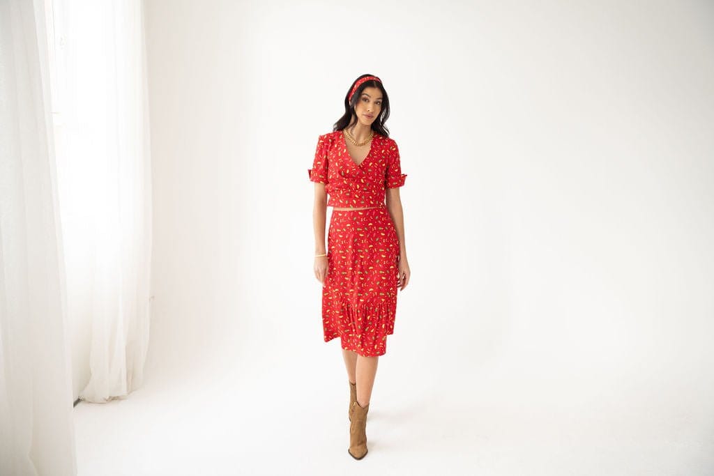 A woman stands in a bright, white room wearing the Atelier Ferrari+Monti Gelato Print Wrap Top, paired with a matching headband and brown ankle boots. She gazes forward with a relaxed expression, hands at her sides, as light filters through sheer curtains on the left.