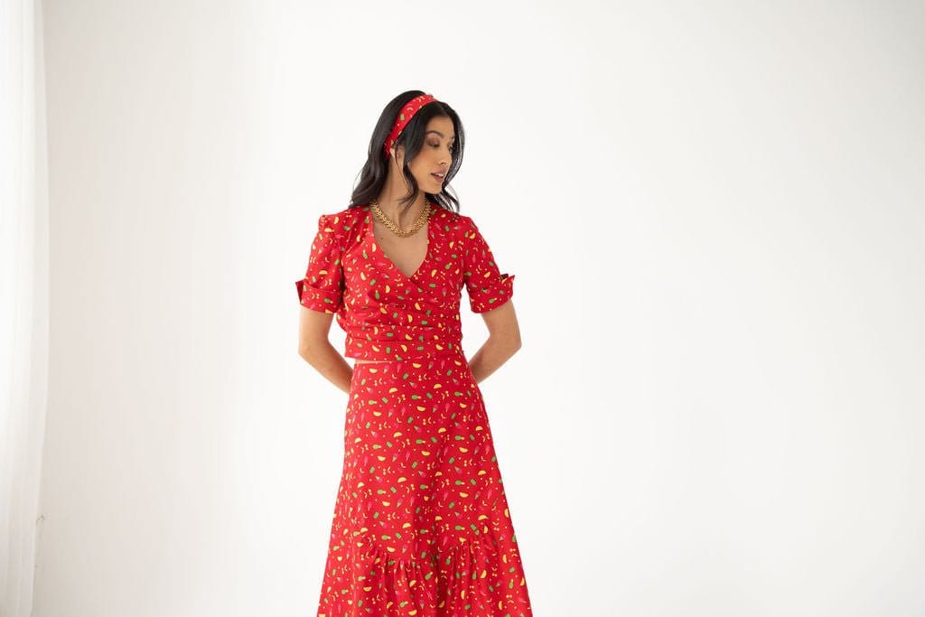 A woman wearing the Atelier Ferrari+Monti Gelato Print Wrap Top, featuring a vibrant red hue with yellow floral patterns, and a matching headband stands against a plain white background, looking down to her left with her hands behind her back.