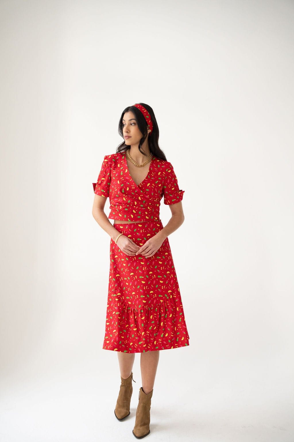 A woman stands against a white background, wearing the Gelato Print Wrap Top by Atelier Ferrari+Monti—an effortlessly charming red wrap top featuring a vibrant floral pattern, short sleeves, and a flattering V-neckline. Her look is finished with a coordinating headband and brown ankle boots as she glances to the side, hands clasped together.