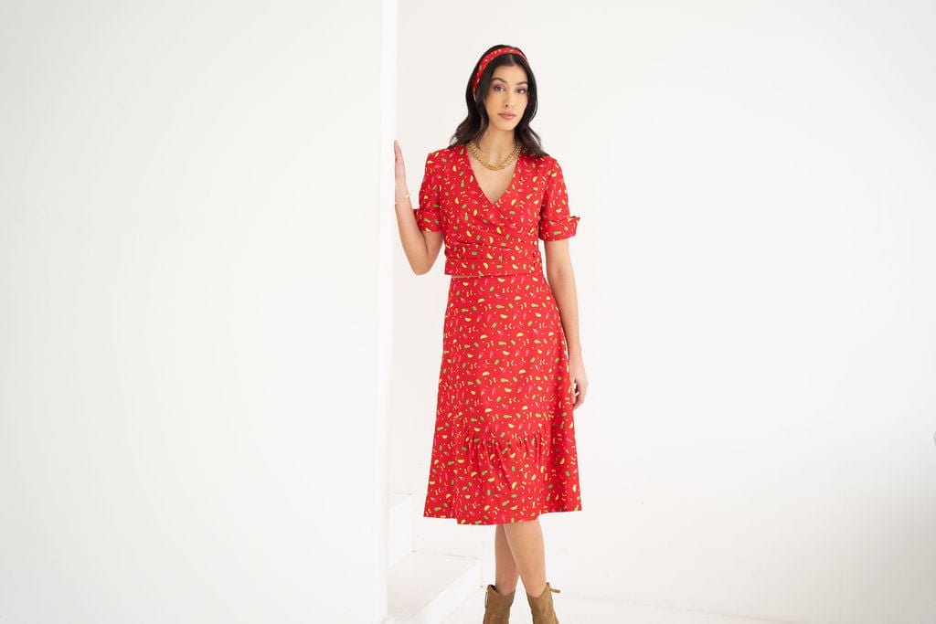 A woman stands in a white room wearing the Atelier Ferrari+Monti Gelato Print Wrap Top with a matching headband, paired with brown ankle boots, and rests one hand on a white wall.