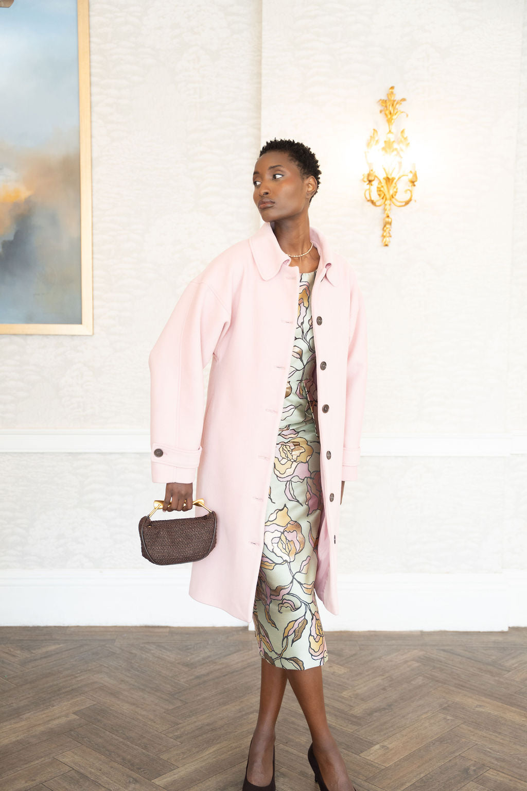 Pastel pink long wool coat with buttons, worn over a patterned midi dress and paired with a small brown handbag.