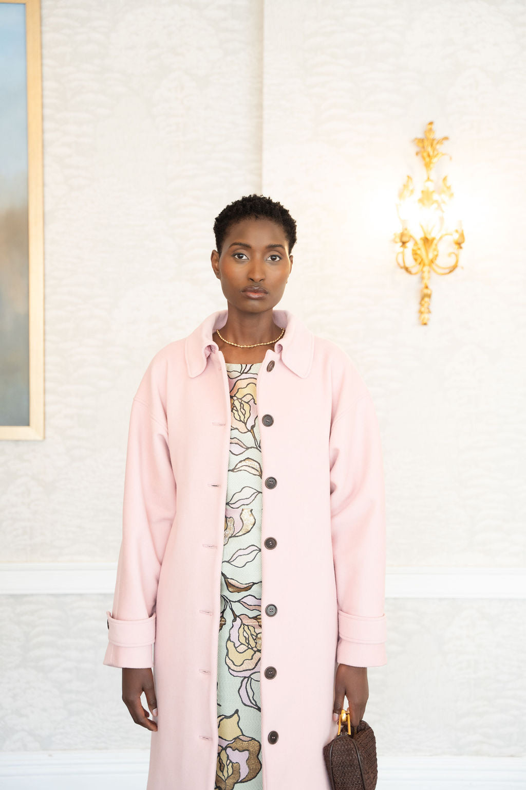 Pastel pink long wool coat with black buttons, worn over patterned dress, held tan handbag.
