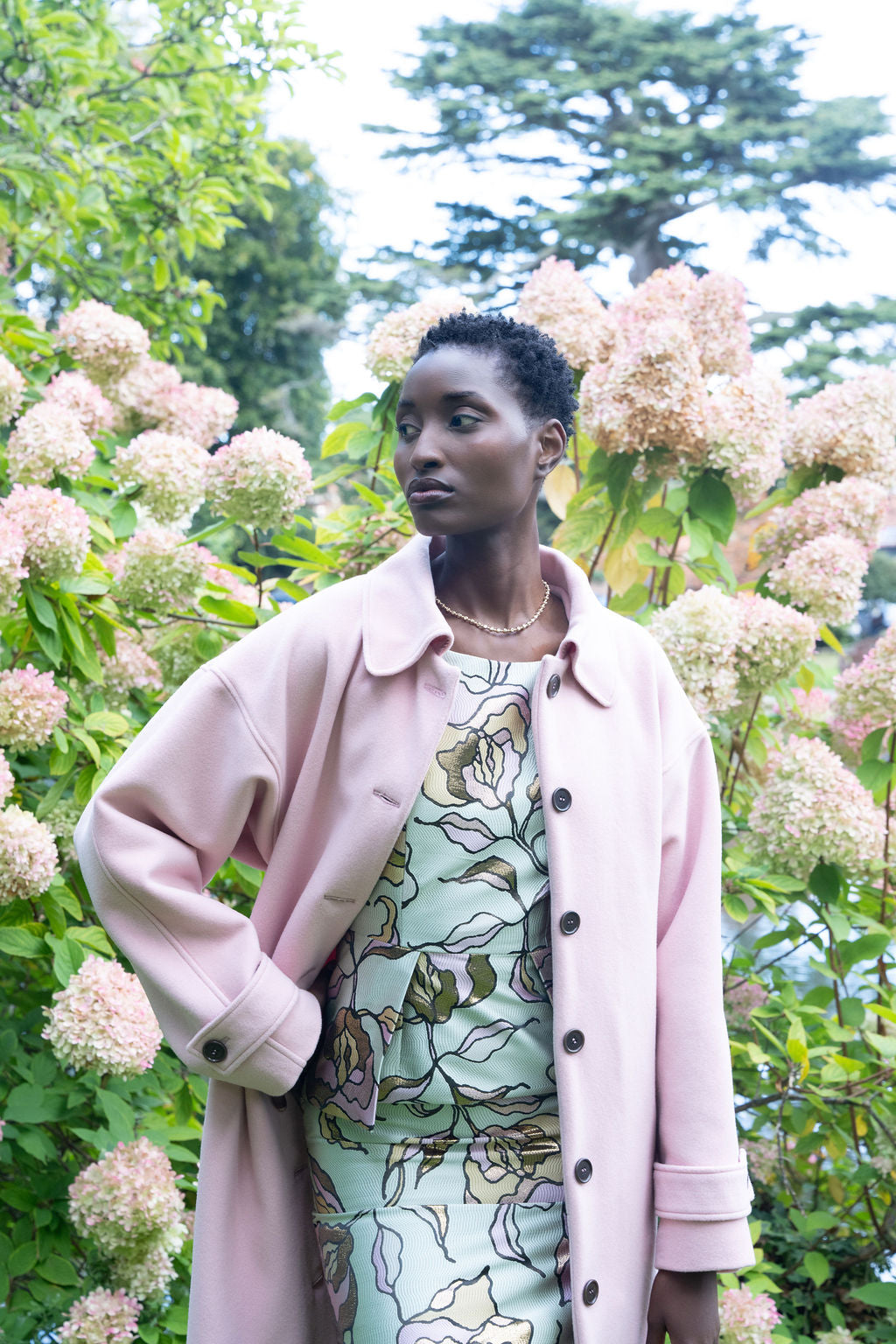 Pastel pink long wool coat with button closure worn over floral print dress outdoors.