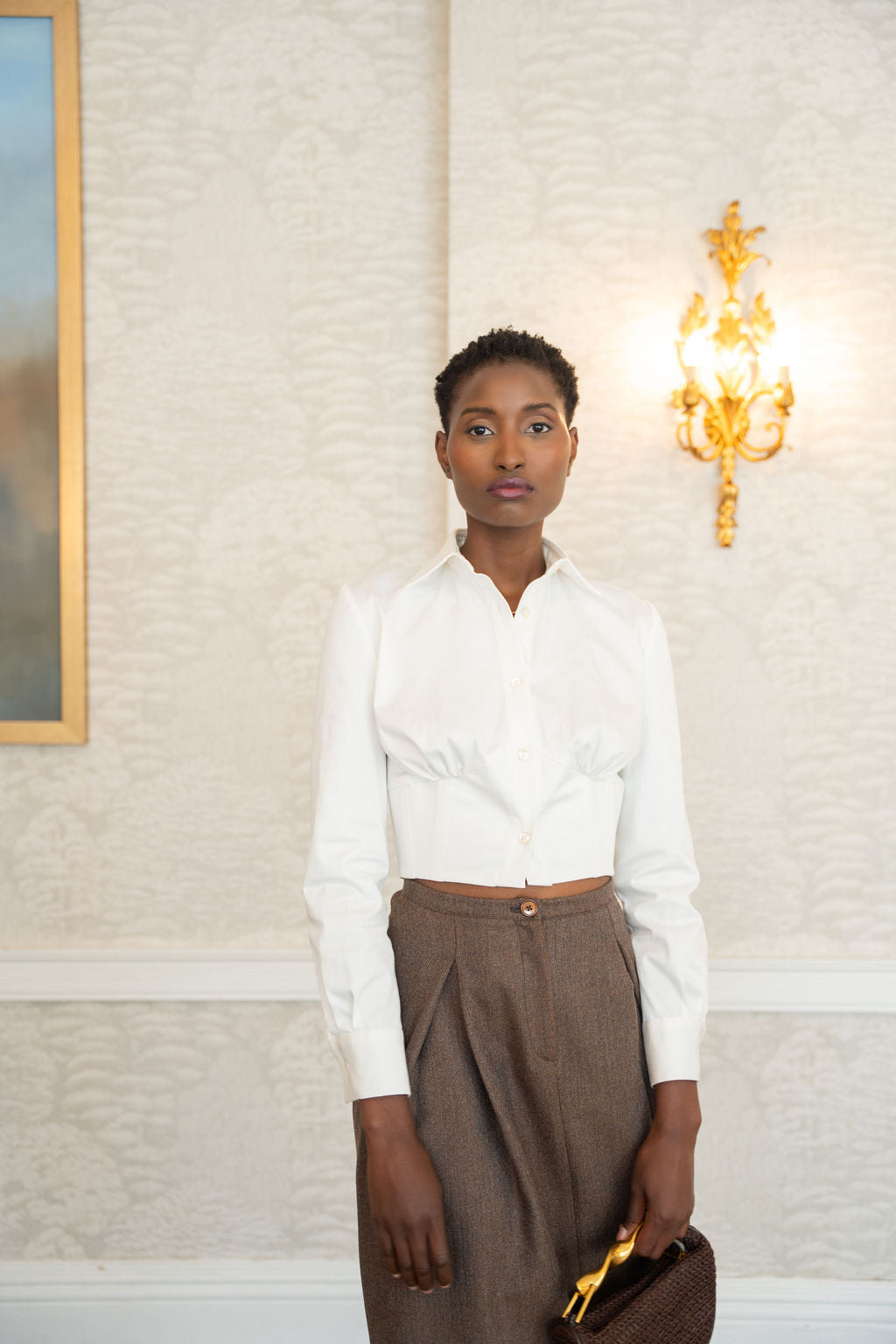 Ivory cotton corset shirt with long sleeves and fitted waist paired with brown trousers.