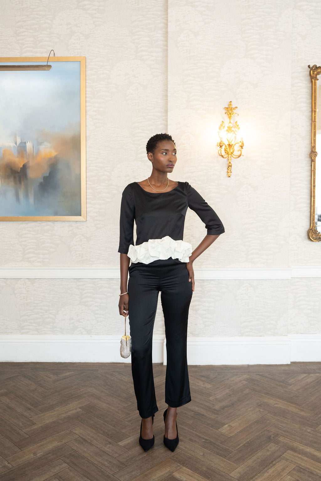 Woman in black outfit with white ruffled top standing in a room with white walls and a painting.