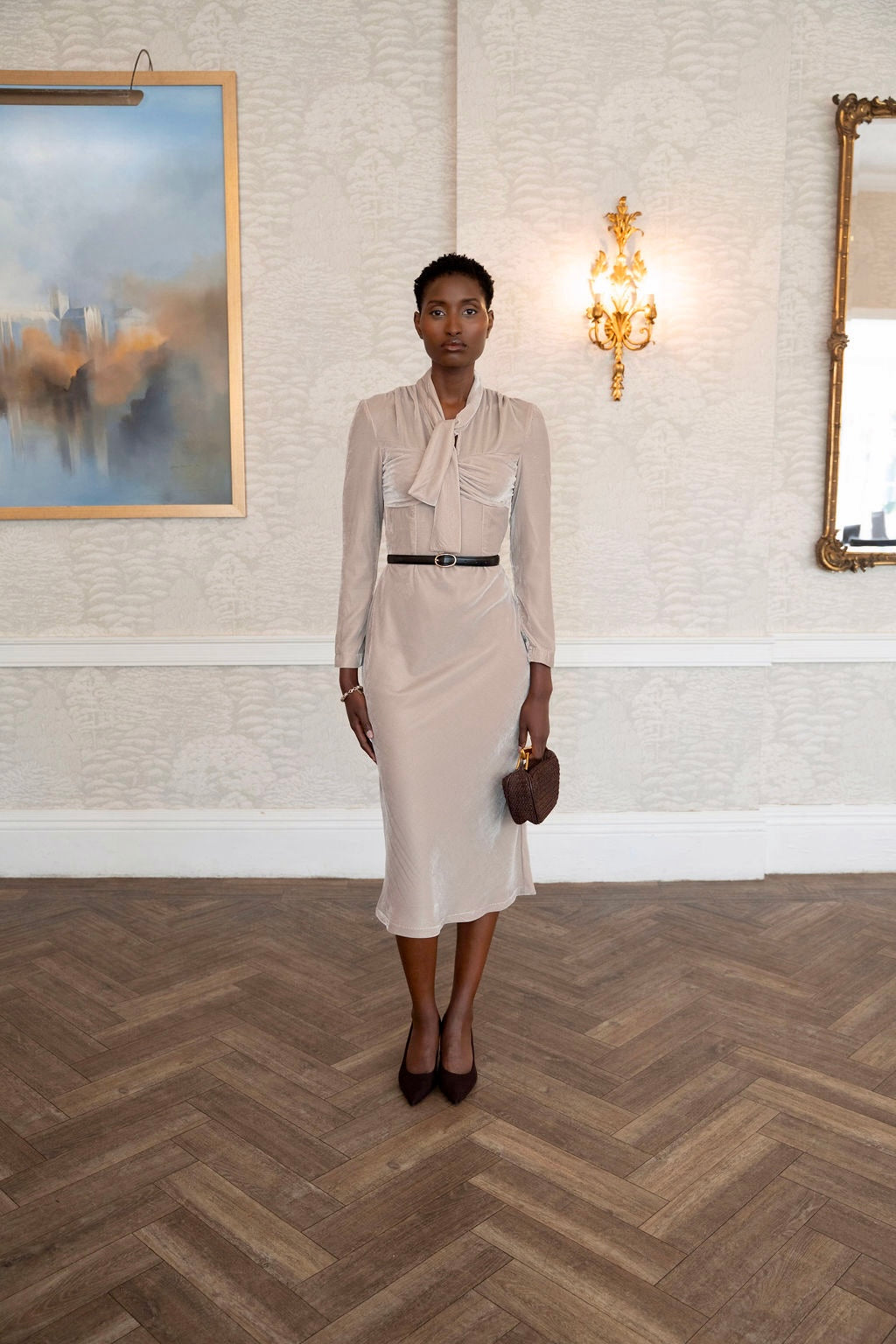 Woman in a elegant velvet beige dress standing in a room with a painting and mirror on the wall
