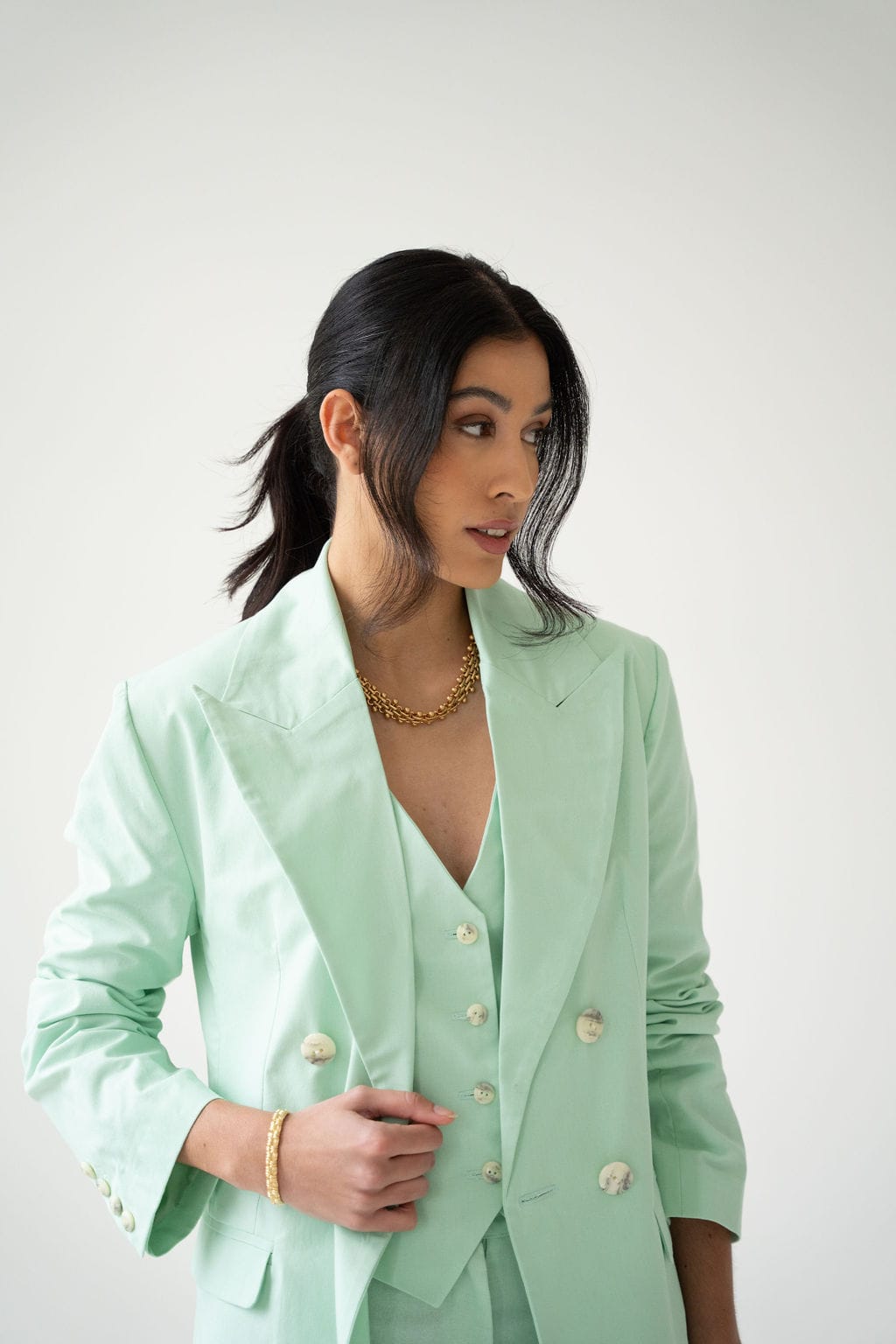 A woman with long dark hair styled in a low ponytail stands against a plain white background, looking to the side. She wears the Atelier Ferrari+Monti Double-Breasted Blazer in mint green cotton, paired with a matching vest and gold jewelry.