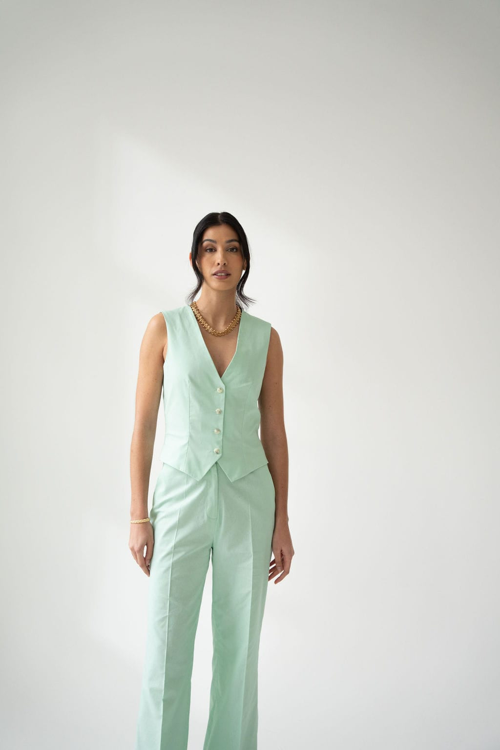 A woman stands against a plain white background, wearing Atelier Ferrari+Monti’s Wide-Leg Tailored Trousers in light mint green with a high-rise waist, paired with a matching sleeveless vest and accessorized with gold jewelry.