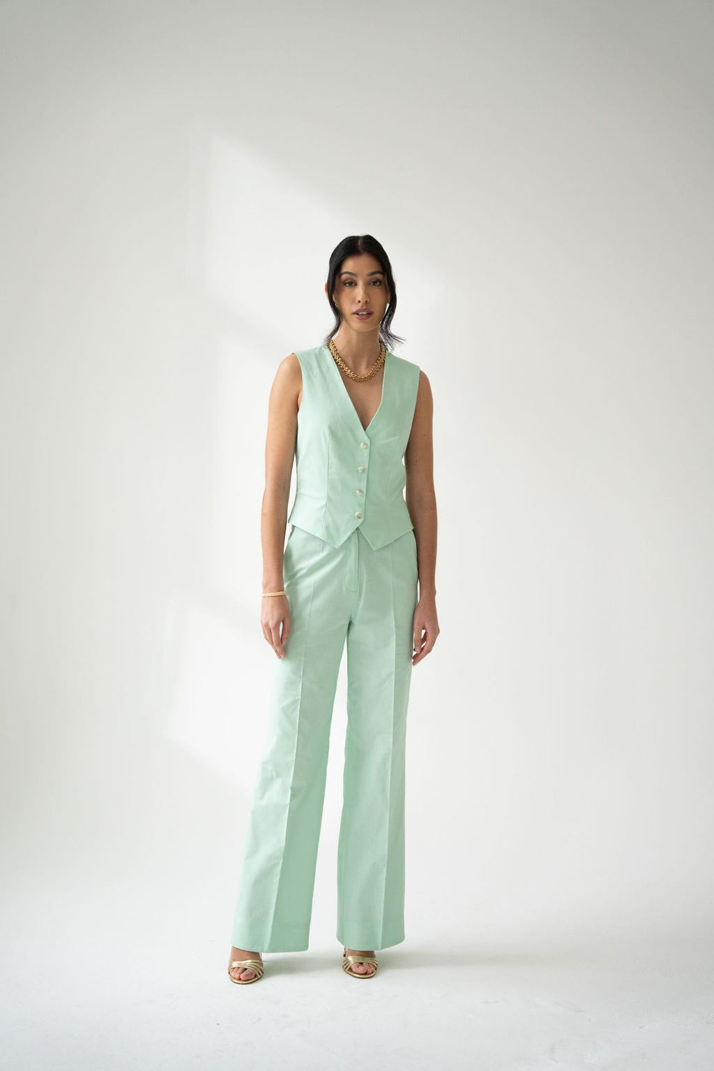A woman wears Atelier Ferrari+Monti’s Wide-Leg Tailored Trousers in mint green cotton, styled with a matching sleeveless buttoned vest, gold sandals, and layered necklaces, set against a plain light background.