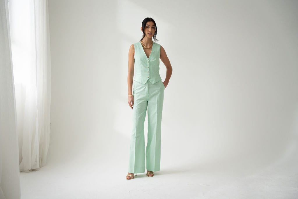A woman stands in a light-filled room wearing Atelier Ferrari+Monti Wide-Leg Tailored Trousers and a matching mint green cotton vest, styled with open-toed shoes and simple jewelry. The background is plain white with sheer curtains on the left.