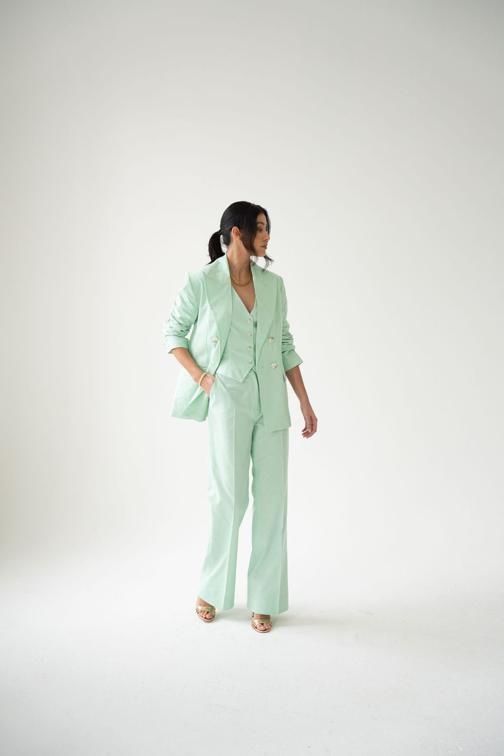 A woman stands against a white background wearing Atelier Ferrari+Monti’s Double-Breasted Blazer in mint green cotton, paired with flared pants and a coordinating vest to showcase elegant women’s suiting. Her long dark hair is tied back, and she gazes to her left with one hand in her pocket.