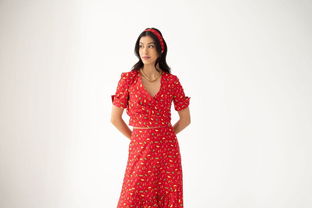 A woman with wavy dark hair stands against a plain white background, wearing the Atelier Ferrari+Monti Gelato Print Wrap Top paired with a matching floral skirt, a coordinating headband, and layered gold necklaces.