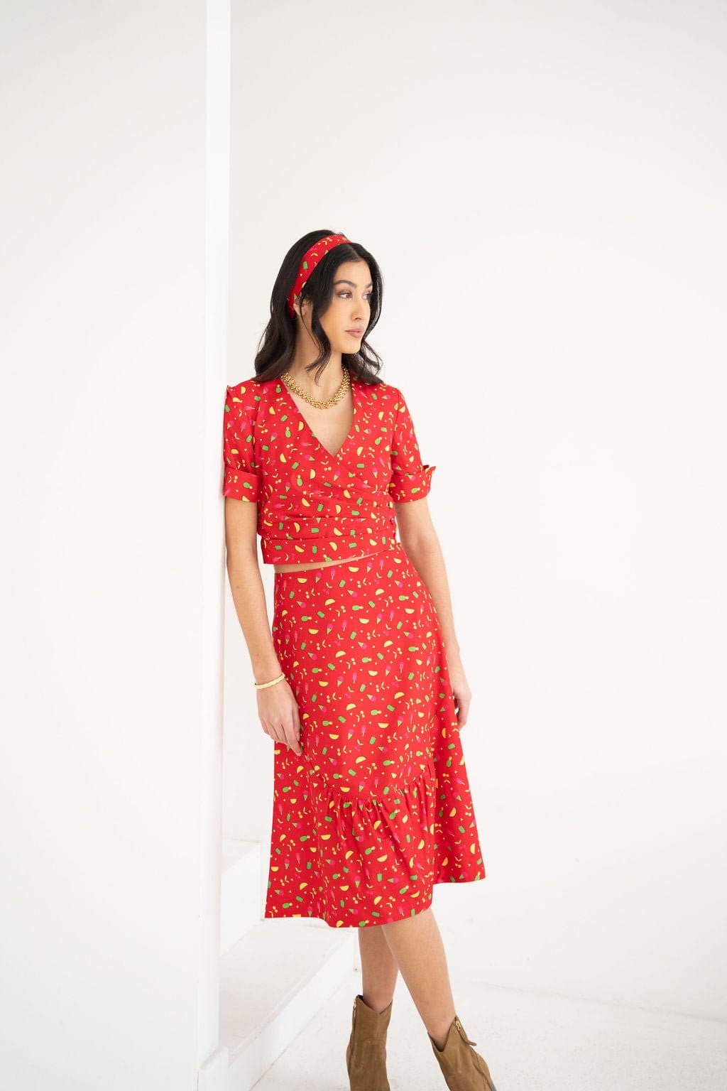 A woman stands against a white wall, looking to the side, dressed in the Atelier Ferrari+Monti Gelato Print Wrap Top paired with a matching red patterned skirt and coordinating headband. She completes her look with brown ankle boots.